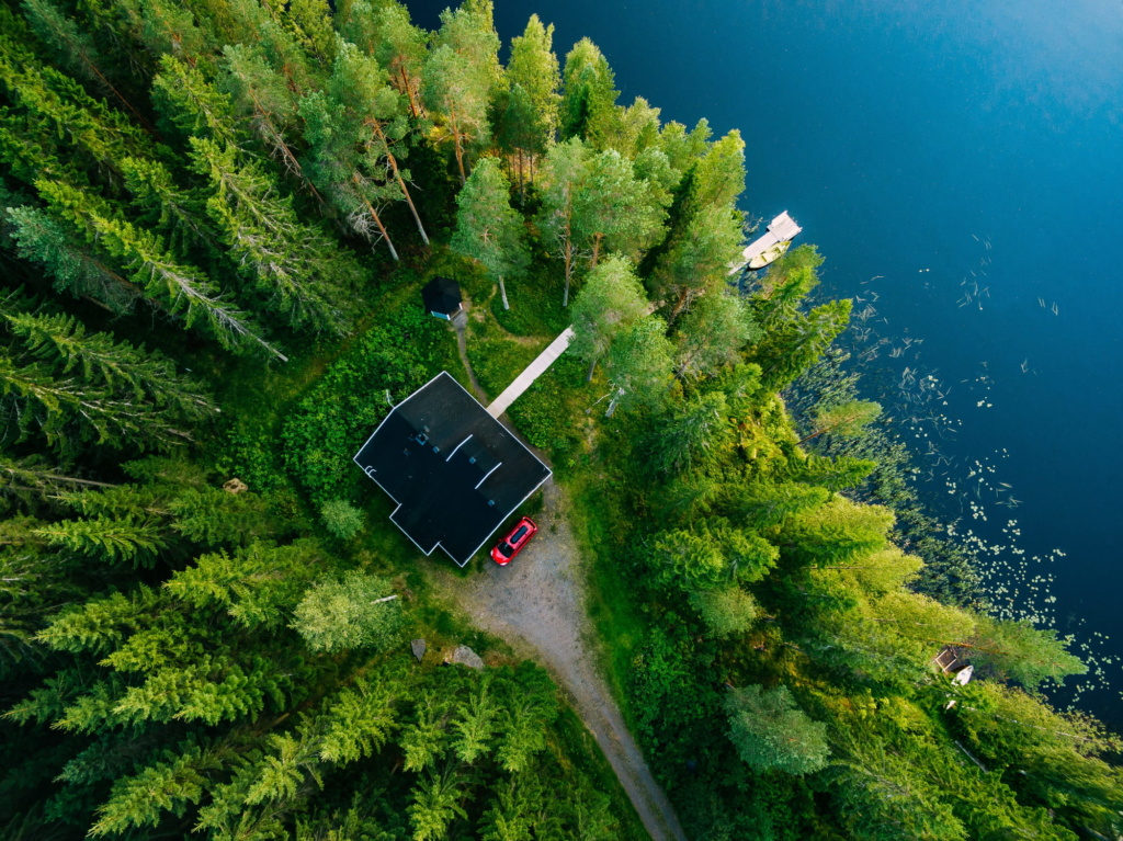 Dronebilde av hytte i grønn skog ved innsjø med brygge. Foto