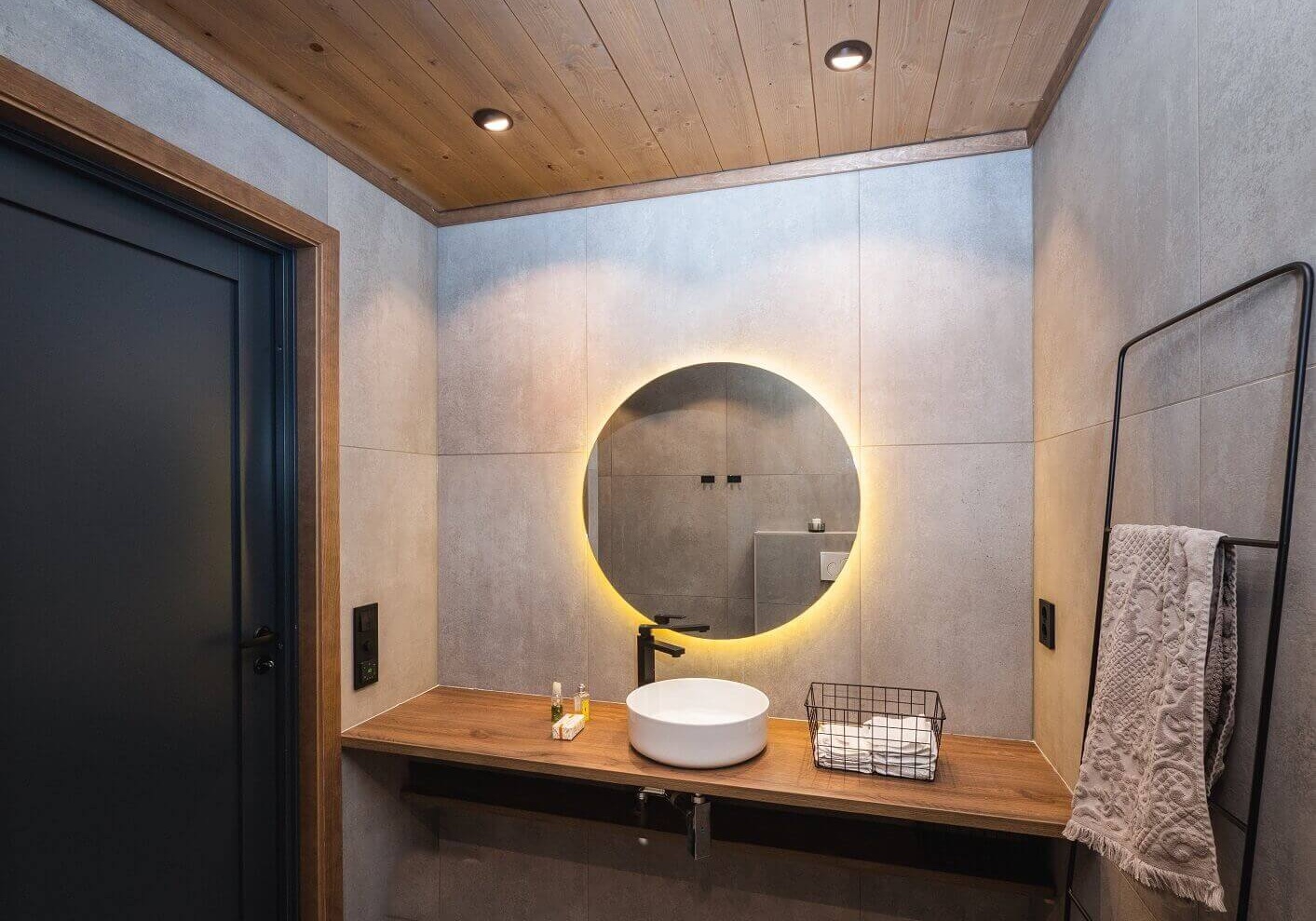 Bad med store grå fliser på vegger. Hvit rund servant og rundt speil med belysning bak. Sorte interiørdetaljer. Foto