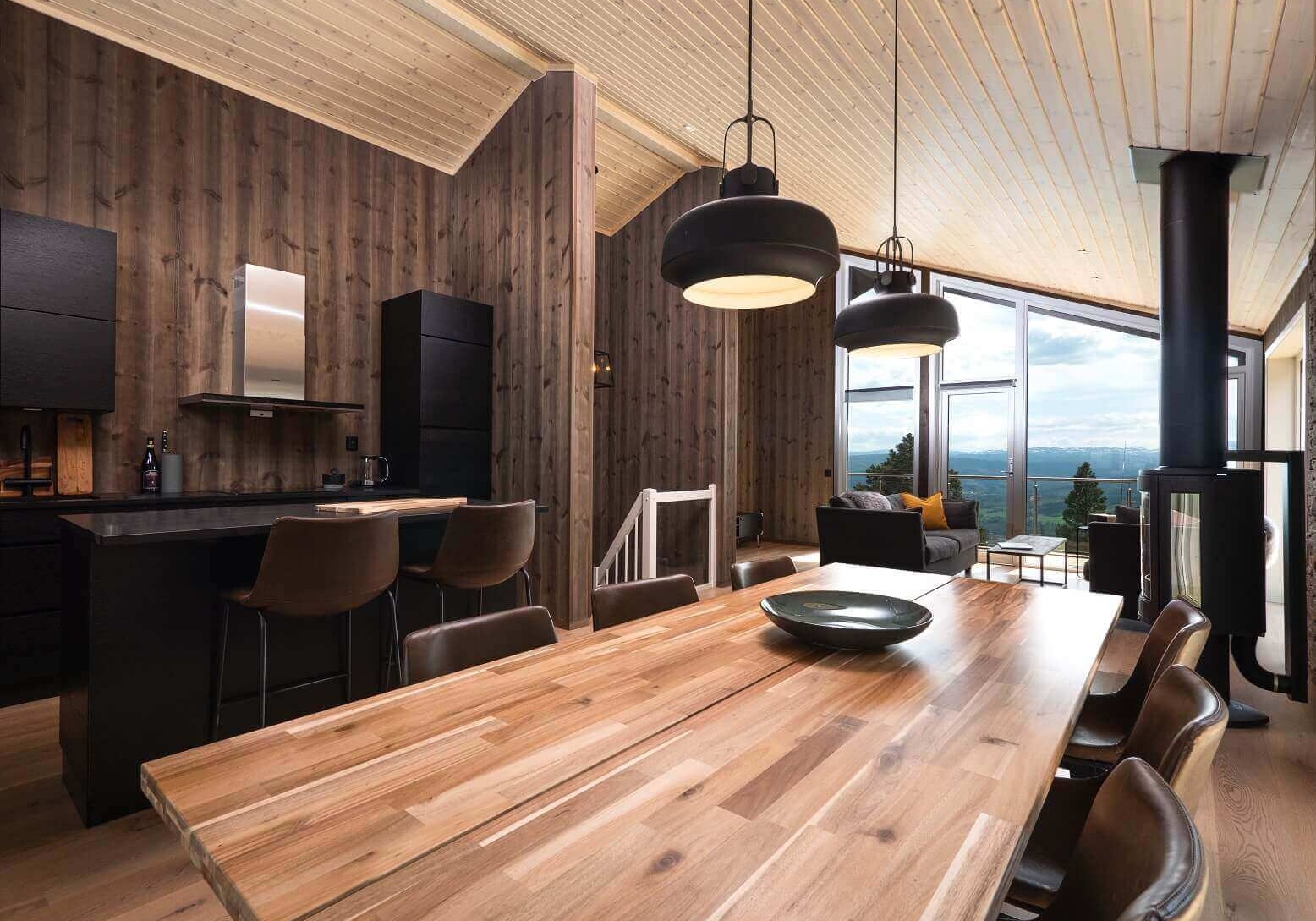 Vindvinkelbilde fra spisestue med kjøkken og stue i bakgrunnen. Brune panelvegger, sort kjøkkeninnredning og trefarget spisebord med stoler i brunt skinn. Foto