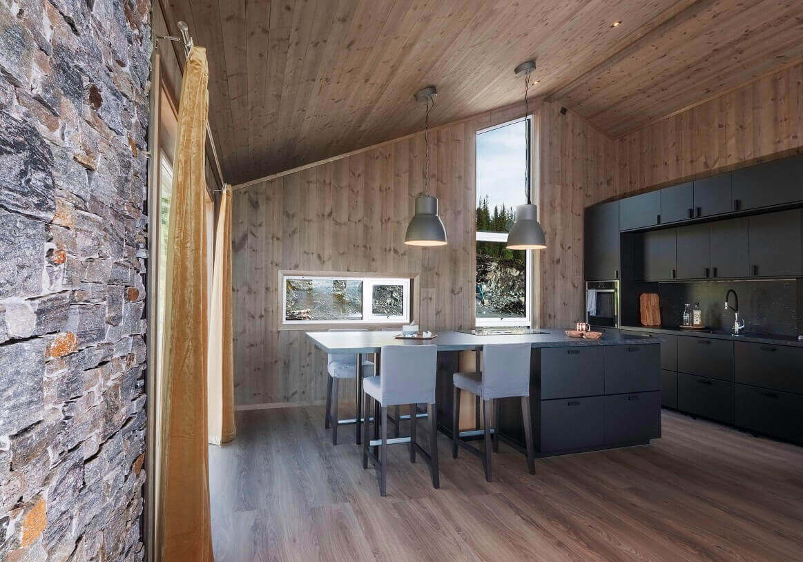 Sort hyttekjøkken med barstoler i sort og grå rundt kjøkkenøy. Brunt panel på vegger og tak. Foto