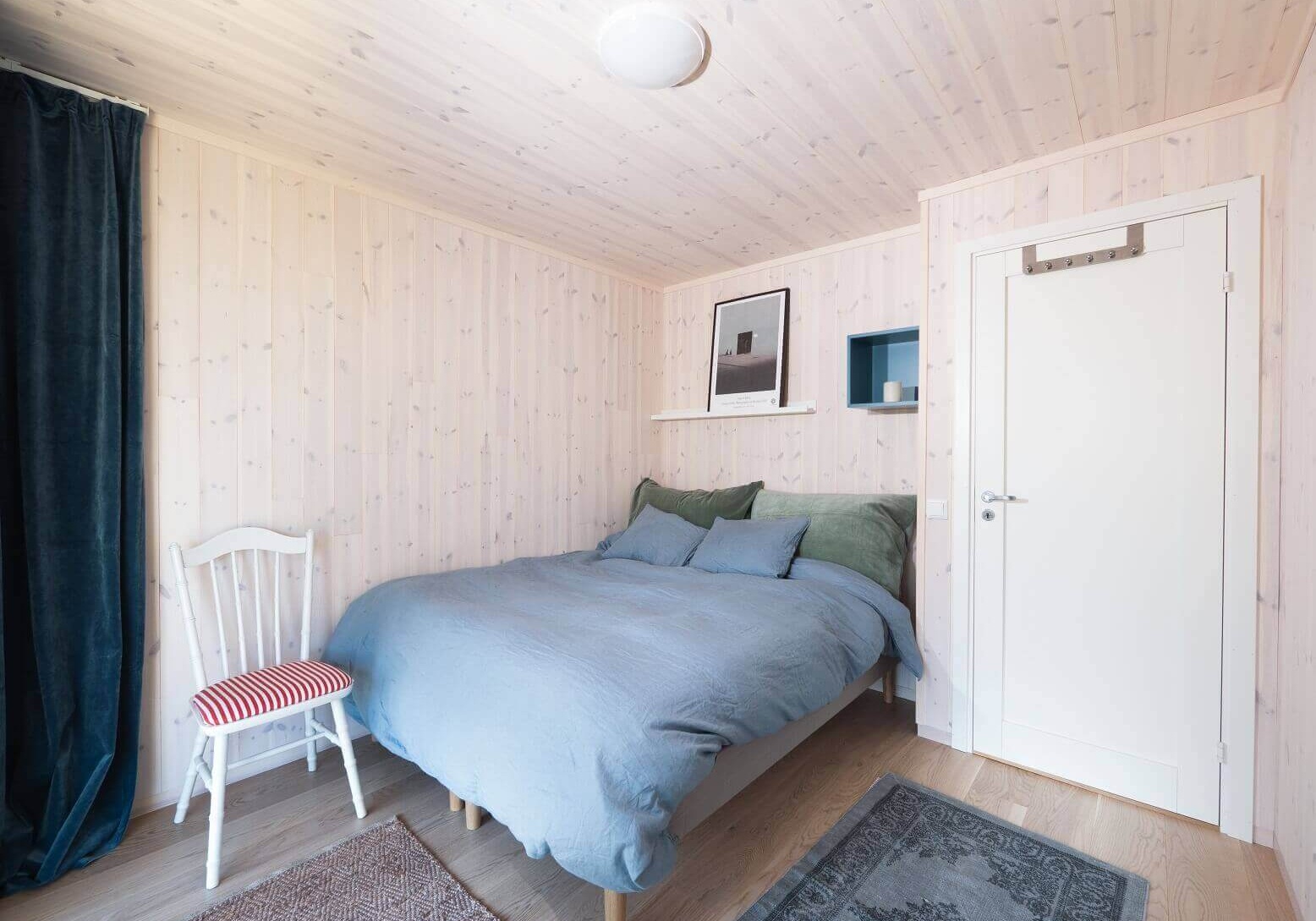 Soverom med hvitkalket panel på tak og vegger. Seng med grått sengetøy og grønne puter. og en vintage hvit stol bak med en rød og hvitstripet pute. Foto