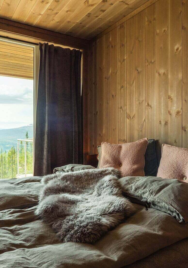 Seng med grått linsengetøy og dusrøde puter. Brune panelvegger bak og vindu med utsikt mot skog. Foto